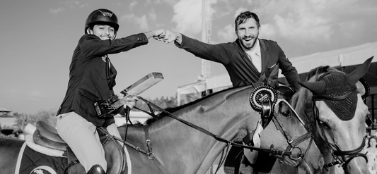 Two equestrians bump fists while mounted on their horses, celebrating a successful show jumping event, exemplifying camaraderie and sportsmanship